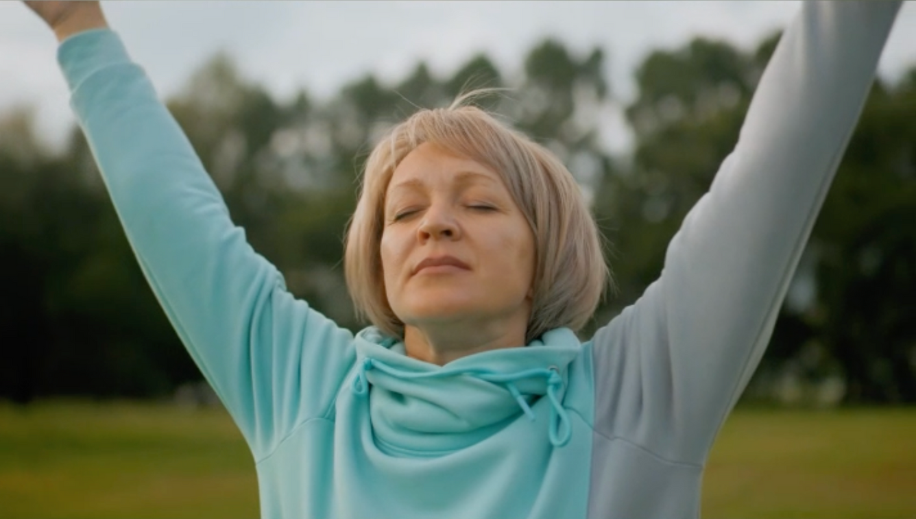 Woman in light blue sweater and scarf raising her arms outdoors